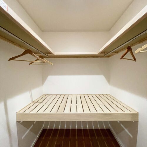 3.7帖のファミリークローゼット。枕棚付きパイプハンガーが両脇の壁にあり、奥には中段と枕棚があります。ハンガーにかけたままの収納と、衣装ケースに入ったものや、お布団なども収納できます。中段、枕棚はすのこタイプ。空気の循環を行い湿気がこもりにくくなります。また、無垢の木材が持つ自然の調湿作用により、湿っぽくなりにくい環境にコントロールしていきます。